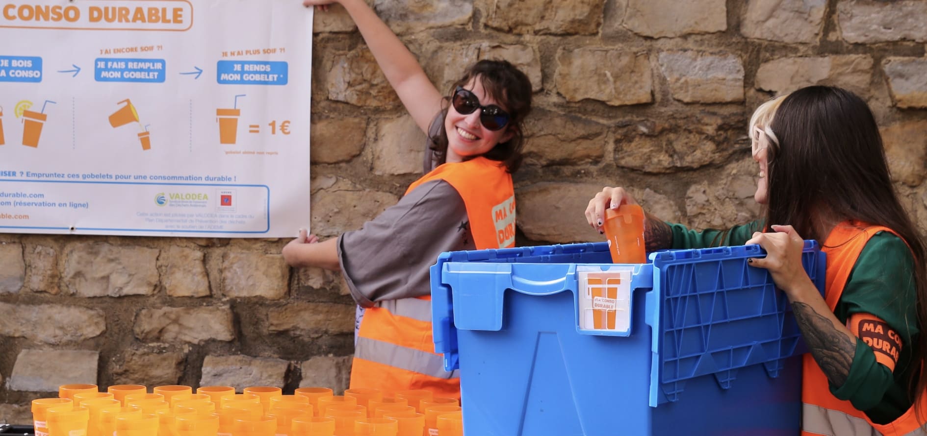 Deux bénévoles en gilet orange devant l’affiche Ma Conso Durable, une caisse de gobelets à la main